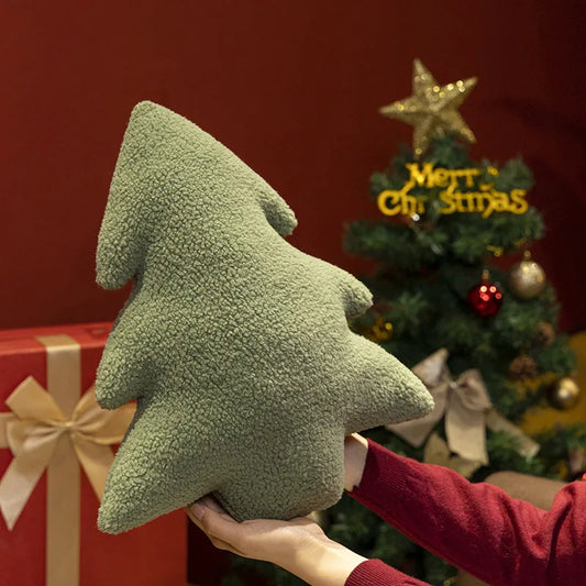 Green Christmas tree-shaped pillow held by a person in front of a decorated Christmas tree.