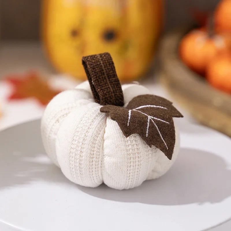 White knitted pumpkin with brown leaf on a white surface with blurred pumpkins in the background