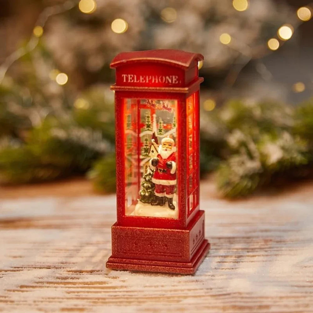 Decorative red telephone booth with a Christmas scene inside, on a wooden surface with blurred greenery in the background.