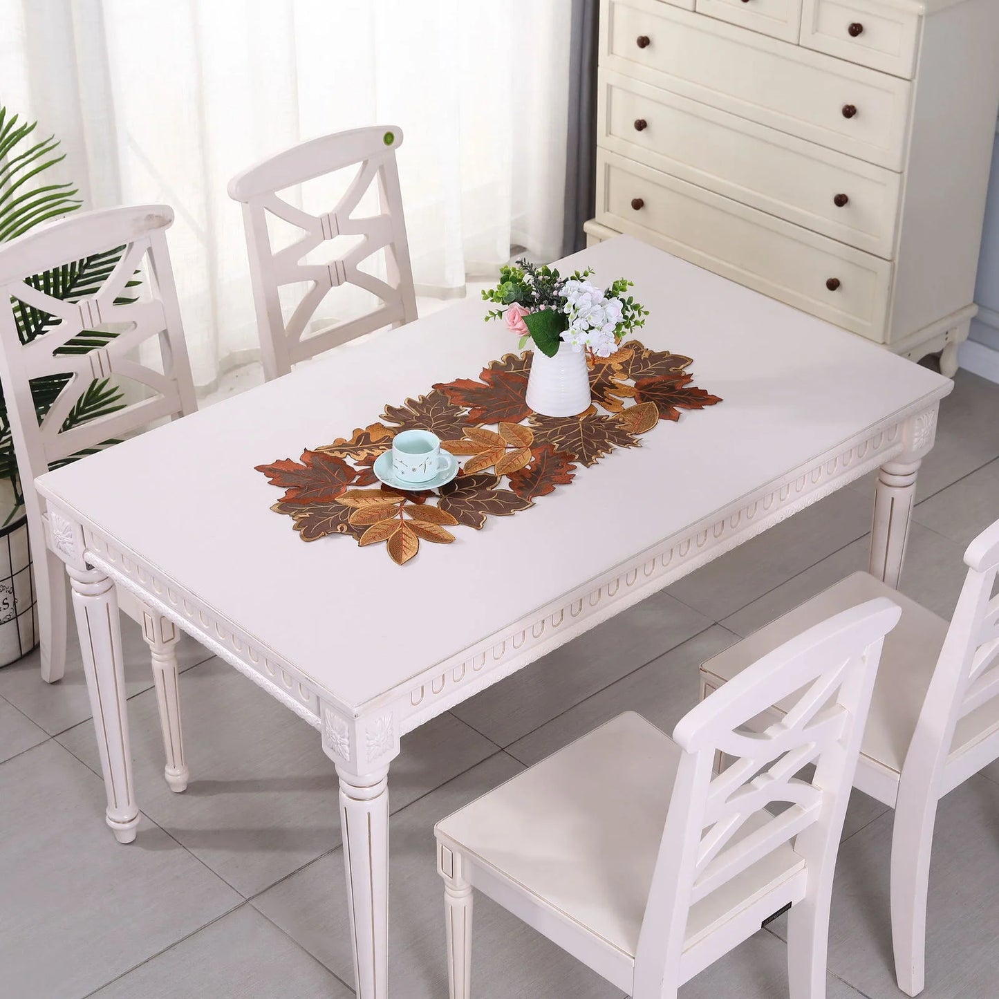 White dining table with four chairs and a decorative leaf runner in a room.