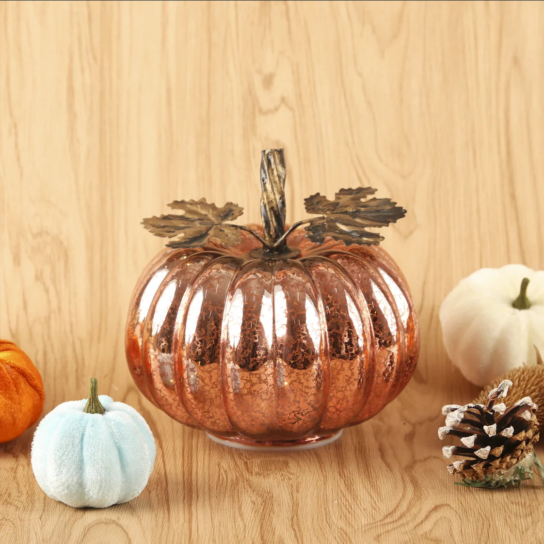 Decorative copper pumpkin on a wooden surface with other pumpkins and a pine cone.