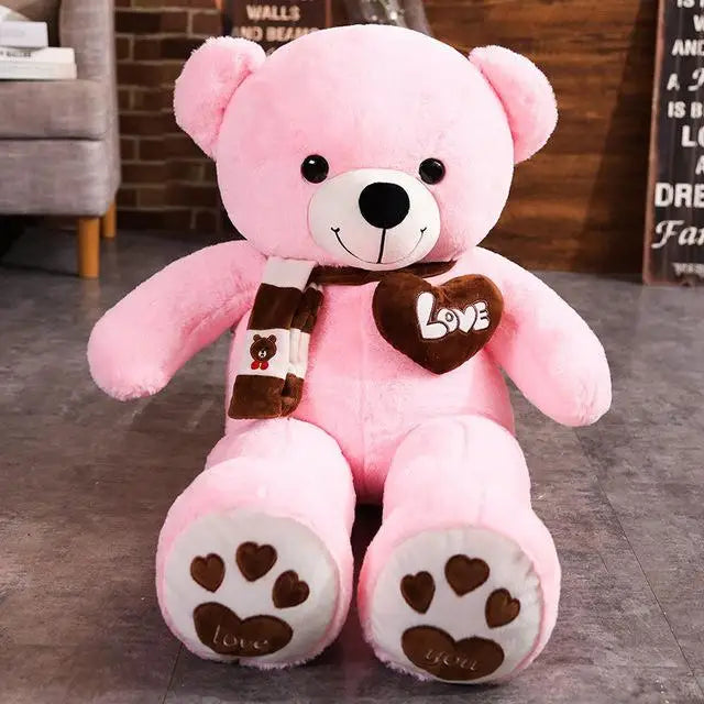 Pink teddy bear with a brown 'LOVE' heart and scarf on a gray floor.
