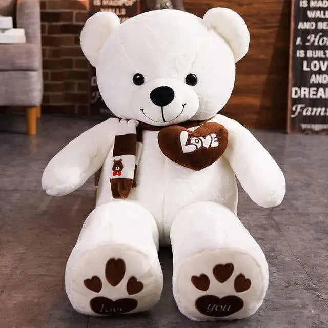 White teddy bear with a brown 'I love you' heart and scarf on a wooden floor.