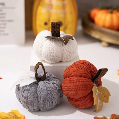 Knitted pumpkins in white, gray, and orange on a light surface with blurred pumpkins in the background.