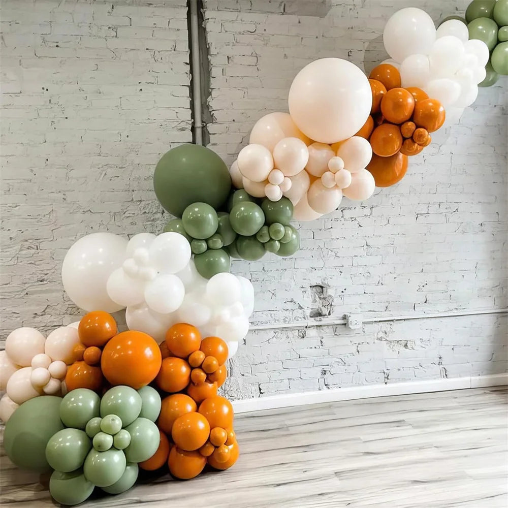 Balloon arch with green, orange, and white balloons against a white brick wall.
