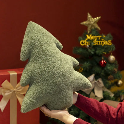 Green Christmas tree-shaped pillow held by a person in front of a decorated Christmas tree.