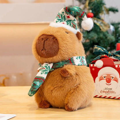 Plush capybara toy with green Christmas hat and scarf in front of a decorated Christmas tree.