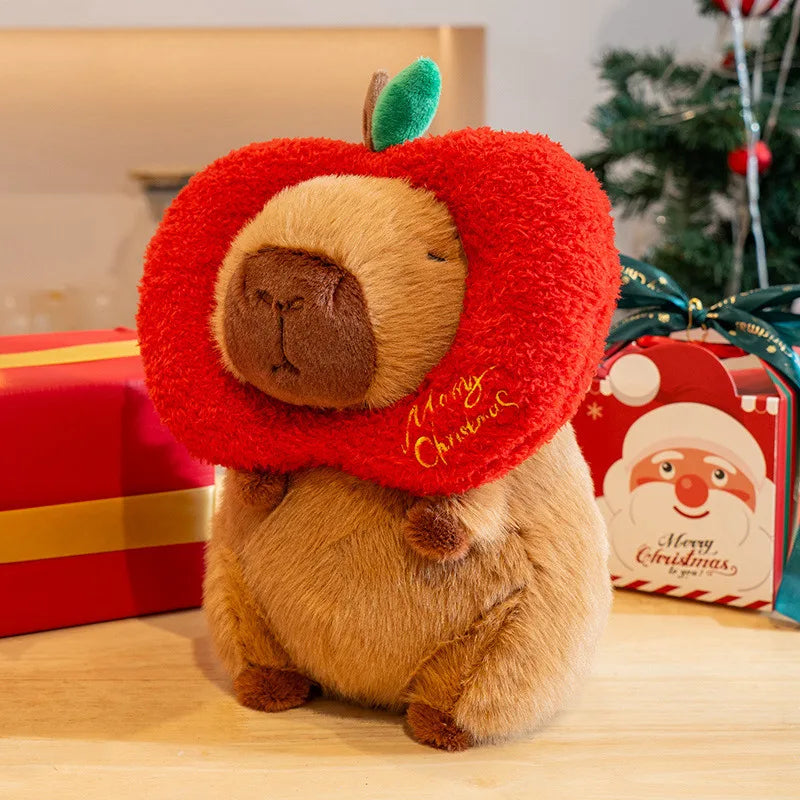 Plush capybara toy wearing a red 'Merry Christmas' apple hat with Christmas presents and decorations in the background.