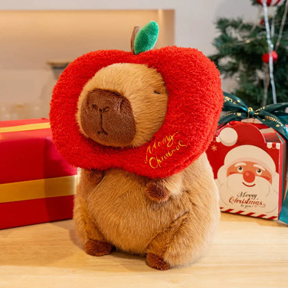 Plush capybara toy wearing a red 'Merry Christmas' apple hat with Christmas presents and decorations in the background.