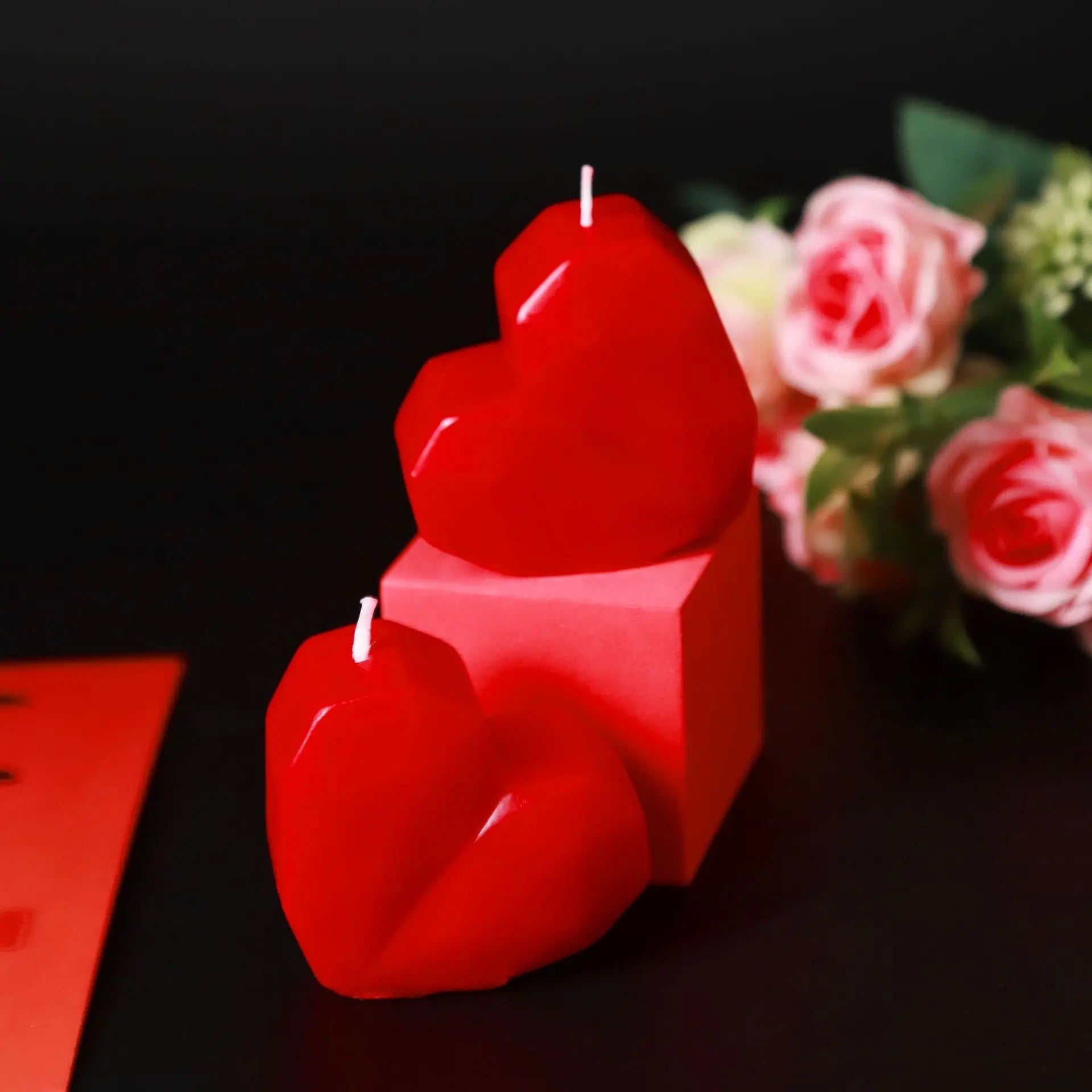 Red heart-shaped candles on a dark surface with pink roses in the background