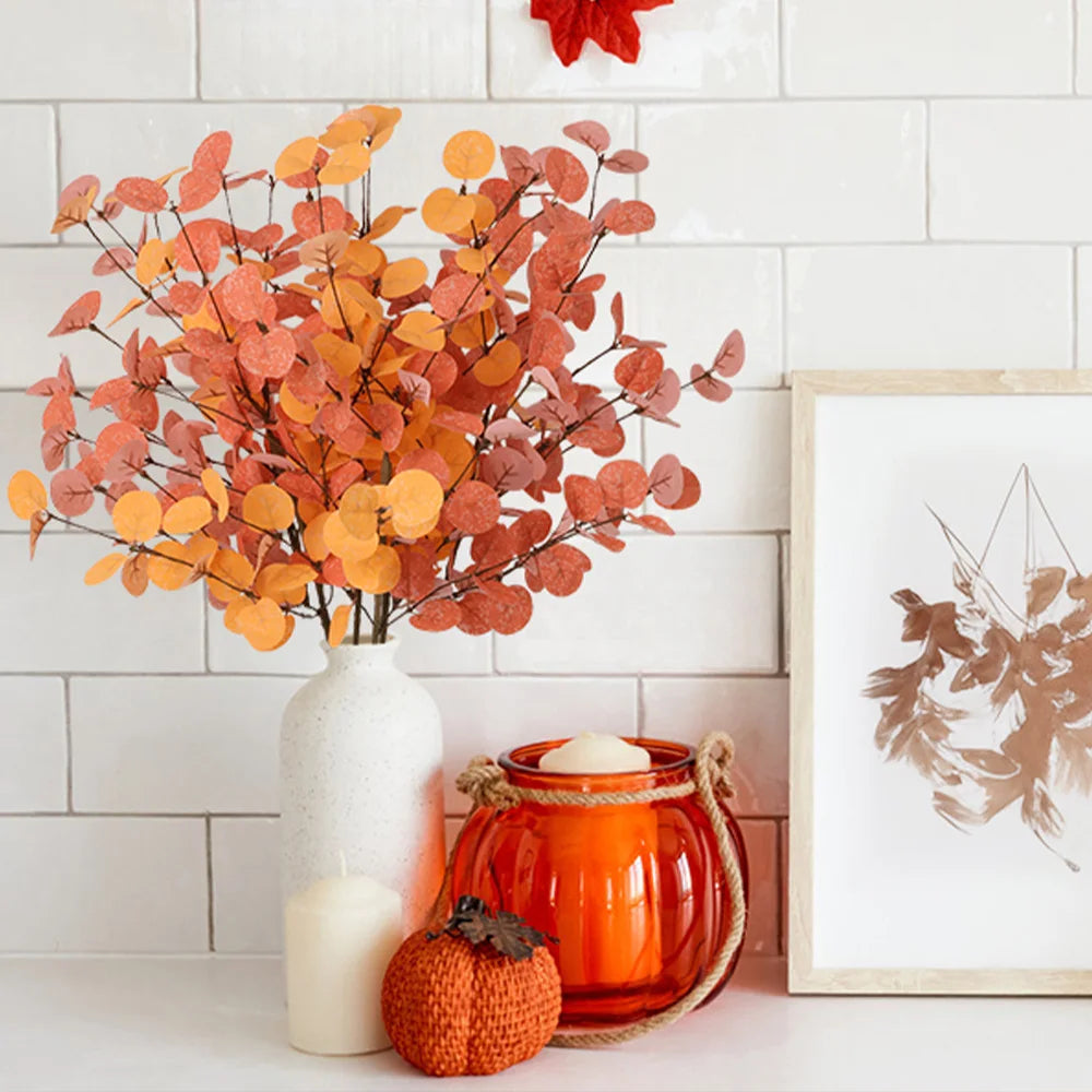 Decorative setup with orange and red leaves in a vase, candle, lantern, and pumpkin against a white tiled wall.