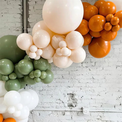 Balloon arch with green, white, and orange balloons against a white brick wall.