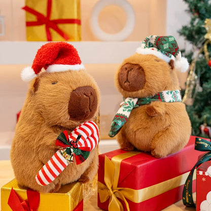 Two capybara teddy bears in festive attire on Christmas presents with a decorated tree in the background.