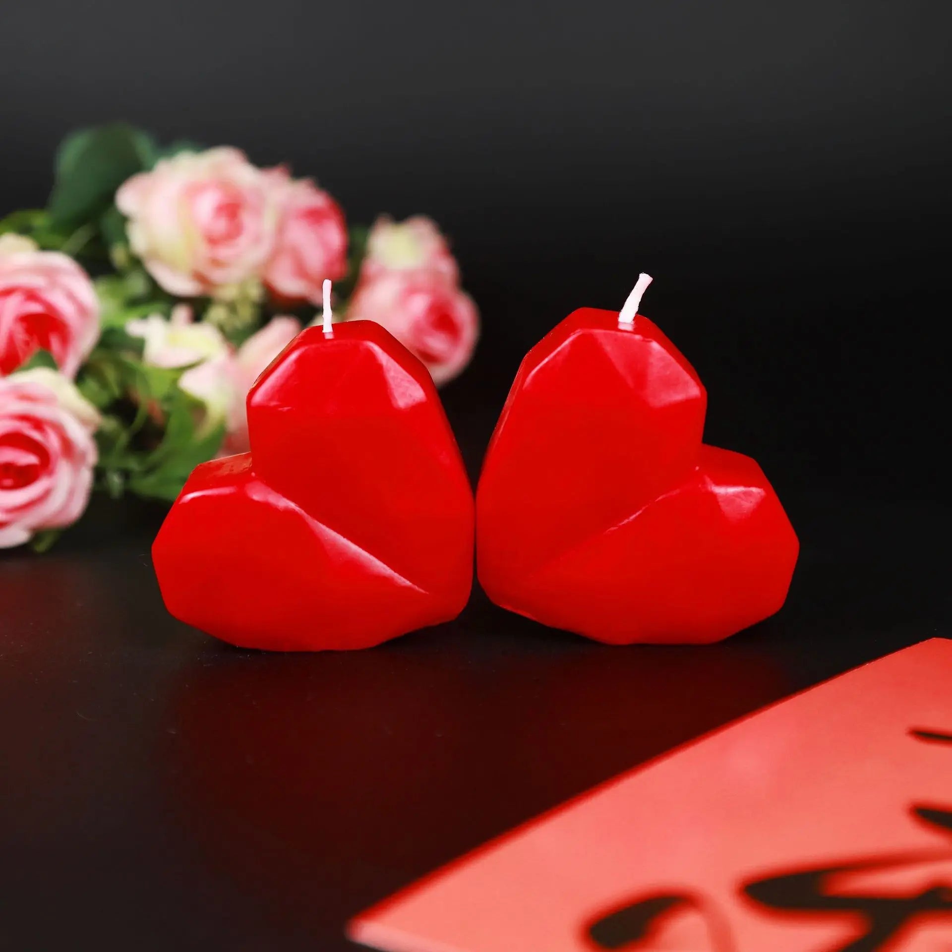 Two red heart-shaped candles on a reflective surface with pink flowers in the background.