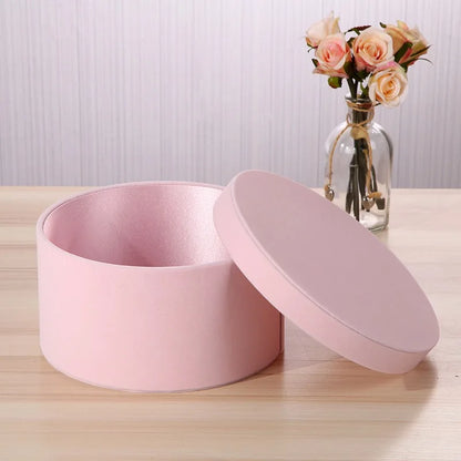 Pink round gift box with a lid on a wooden surface, next to a vase with flowers.