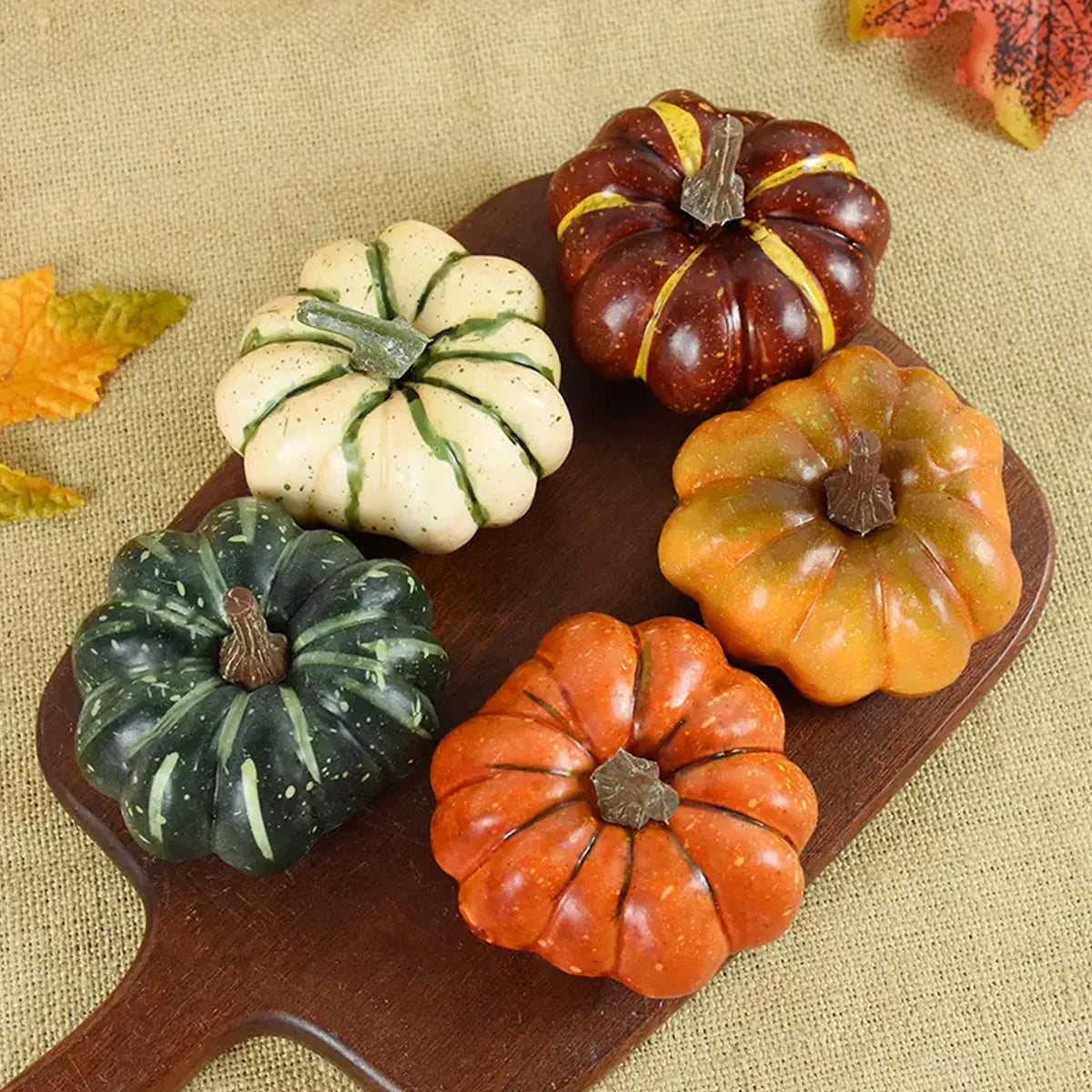 Five small pumpkins in various colors on a wooden board with a beige fabric background.