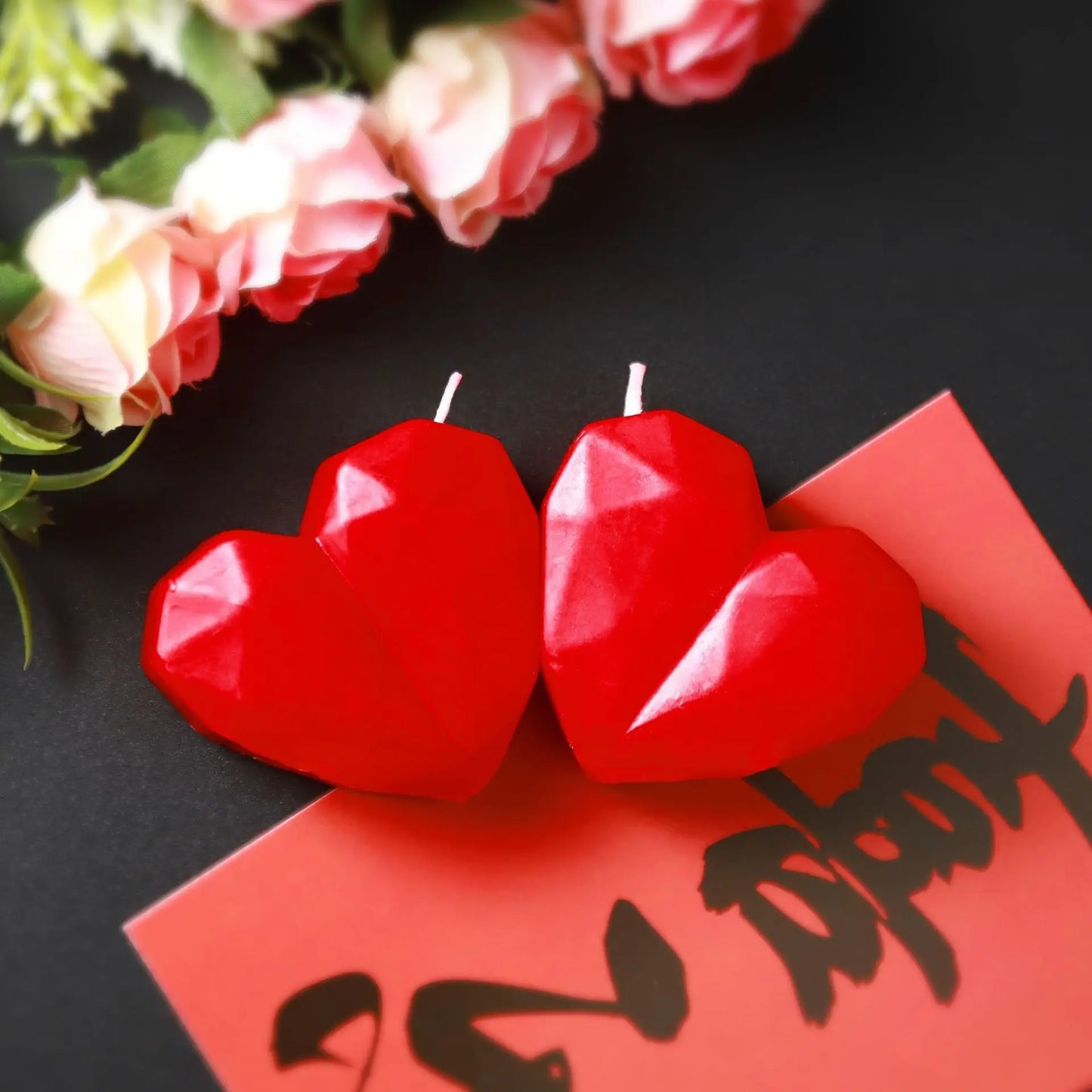Two red heart-shaped candles on a black surface with flowers in the background