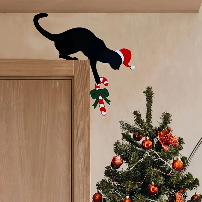 Black cat wearing a Santa hat next to a Christmas tree with decorations on a beige wall.