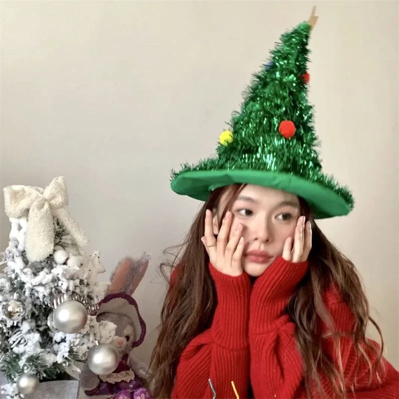 Person wearing a green Christmas tree hat with ornaments, sitting next to a decorated Christmas tree.